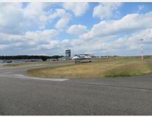 Kleine Flugzeuge auf einem Flugplatz mit Rollfeld und Grasflächen, im Hintergrund ein grauer Kontrollturm und Wald. Der Himmel ist teils bewölkt mit blauem Hintergrund.