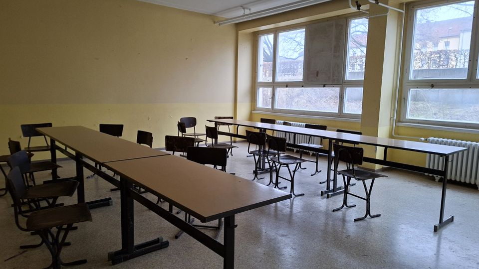 Leerer Klassen- oder Seminarrraum mit mehreren langen Tischen und einzelnen Stühlen, beige gestrichenen Wänden und linoleumbedecktem Boden. Große Fenster entlang einer Wand lassen Tageslicht herein; darunter stehen Heizkörper und sichtbare Rohrleitungen verlaufen an Decke und Wand.