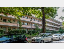 Wohngebäude mit drei Etagen in pastellorange Farbe und umlaufenden Balkonen, umgeben von Bäumen und gepflegten Grünflächen; im Vordergrund geparkte Autos auf einer gepflasterten Straße.