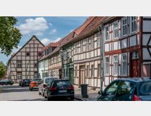 Eine Straße mit historischen Fachwerkhäusern, die sich in einer Reihe aneinanderreihen. Geparkte Autos säumen die gepflasterte Straße, und eine Straßenlaterne steht am Rand; die Dächer der Häuser sind mit roten Ziegeln gedeckt.