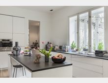 Moderne Küche mit weißen Einbauschränken und einer großen Arbeitsinsel mit dunkler Oberfläche, darauf eine Vase mit rosa Tulpen und eine Schale mit Obst. Ein großes Fenster mit Blick ins Grüne sorgt für viel Tageslicht, Pflanzen stehen auf der Fensterbank.