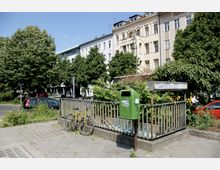 Straßenansicht mit einem gelben Fahrrad an einem Metallgeländer, einem grünen Mülleimer und einem überdachten Treppenzugang, der in eine U-Bahn-Station führt. Im Hintergrund stehen mehrstöckige Wohnhäuser, umgeben von Bäumen und parkenden Autos.