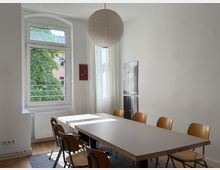 Heller Besprechungs- oder Essraum mit langem rechteckigem Tisch und mehreren Holzstühlen, darüber eine große runde Papier-Hängelampe. Links ein großes Fenster mit Blick auf Bäume und ein gegenüberliegendes Gebäude, an der Wand hängen zwei gerahmte Bilder.