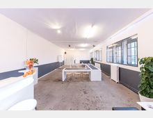 Großer, heller Besprechungsraum mit Betonboden, weißen Wänden, grauem Wandstreifen und rechteckigem Tisch in der Mitte, umgeben von transparenten Stühlen. An der rechten Seite befinden sich große Fenster und ein Heizkörper, links steht eine kleine weiße Theke mit einer Schale Obst.