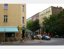 Straßenszene mit einer Eckkneipe namens "Thüringer-Stuben" in einem gelben Gebäude mit grün-weiß gestreifter Markise. Im Hintergrund sind bunte Wohnhäuser mit Balkonen und Bäume zu sehen, während Autos auf der Straße und am Straßenrand geparkt sind.