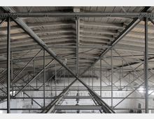 Blick auf das Innere einer Industriehalle mit sichtbarer Dachkonstruktion aus Stahlträgern und Holzplanken sowie weißen Ziegelwänden im Hintergrund. Die Struktur zeigt diagonale Verstrebungen und Hängelampen an den Trägern.