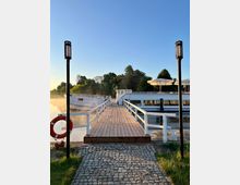 Holzsteg mit Geländer, der zu einem festgemachten Schiff führt, flankiert von zwei Laternen. Im Hintergrund Wasser, Bäume und ein klarer, blauer Himmel.