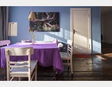 Esszimmer mit Holzboden, einem Tisch mit lila Tischdecke und weißen Holzstühlen. An der blauen Wand hängt ein großformatiges Naturbild, daneben ein weißer Türrahmen, durch den Sonnenlicht fällt.