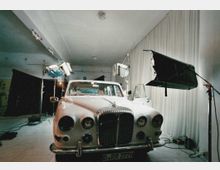 Ein weißer Oldtimer mit geöffneten Türen wird in einem Studio mit professionellen Beleuchtungsgeräten fotografiert. Der Raum hat weiße Wände, einen gefliesten Boden und einen Vorhang im Hintergrund.