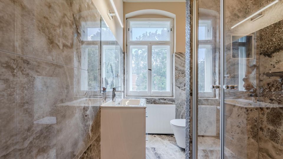 Modernes, schmales Badezimmer mit großformatigen marmorierten Wand- und Bodenfliesen, Waschtisch und Spiegel an der linken Wand sowie einer verglasten Dusche rechts. Am Ende des Raums befindet sich ein großes Fenster mit Oberlicht über einem Heizkörper, daneben ein WC.