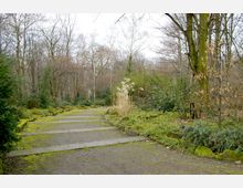 Ein schmaler Weg aus Steinplatten führt durch einen parkähnlichen Wald mit kahlen Bäumen und grünem Unterbewuchs. Links und rechts des Weges befinden sich moosbedeckte Steine und Büsche, während der Hintergrund von dichtem Gehölz dominiert wird.
