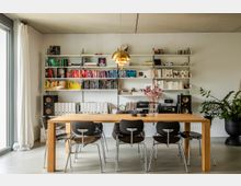 Wohnzimmer mit modernen Möbeln: Ein großer Holztisch, umgeben von schwarzen Stühlen, steht vor einer Wand mit weißen Bücherregalen, die Bücher, Aktenordner und Dekorationsgegenstände enthalten. Im Raum befinden sich außerdem eine goldene Hängelampe, Lautsprecher, eine Stereoanlage und eine große Zimmerpflanze.