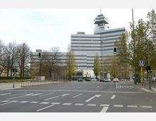 Breite Straßenkreuzung mit markierten Fahrspuren und Verkehrsampeln, im Hintergrund ein modernes Bürogebäude mit grauen horizontalen Fassadenstreifen und einem markanten Turmaufbau. Umgeben von Bäumen, die teilweise entlaubt sind, und einer U-Bahn-Station mit blauem Schild und Aufschrift „U2“ am rechten Bildrand.