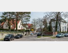 Straßenszene in einem Wohngebiet mit freistehenden Häusern im klassischen Stil, roten Ziegeldächern und Vorgärten, flankiert von Bäumen. Geparkte Autos stehen entlang der Straße, die als Tempo-30-Zone ausgewiesen ist.