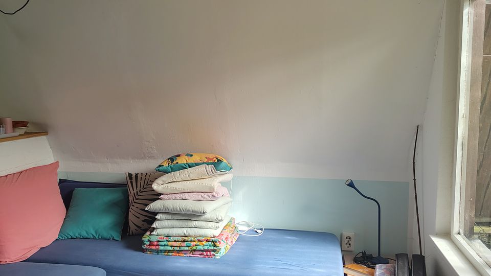 Kleines Zimmer mit schräger weißer Holzdecke und sichtbaren Balken, zwei niedrigen Betten aus Europaletten mit blauen Matratzen und mehreren Kissen. Rechts fällt Tageslicht durch ein großes Fenster; daneben stehen eine kleine Holz-Nachttischablage, eine Leselampe und ein gepolstertes Möbel an der Wand.