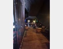 Dunkler Innenraum einer Lounge mit Holzboden und gedämpfter Beleuchtung. Im Hintergrund sind Sitzgruppen mit Sesseln und Tischen sowie mehrere Personen zu sehen, die in einer gemütlichen Atmosphäre zusammensitzen.