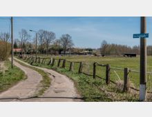 Ein ländlicher Weg mit Betonplatten führt entlang eines Weidezauns, der eine grüne Wiese mit vereinzelten Bäumen und weidenden Pferden umgibt. Im Hintergrund befinden sich ein Gebäude mit rotem Dach, weitere Zäune und eine kleine Holzhütte.