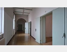Langer, leerer Flur in einem älteren Gebäude mit hohen weißen Wänden, Fensterreihe links und mehreren offenen, hellblauen Türen zu Nebenräumen. Deckenleuchten hängen an grünen Trägern, am Ende des Ganges befindet sich ein Rundbogen mit einer weiteren Tür.