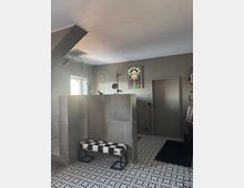 Modernes Badezimmer mit grauen Fliesenwänden und einem gemusterten weißen und schwarzen Boden. In der Mitte des Raums steht eine Bank mit schwarz-weißem Kuhfellmuster, darüber hängt ein farbenfrohes Kunstwerk mit dem Porträt einer Frau an der Wand.