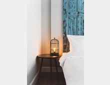 Nachttisch mit dunkler Holzoberfläche, darauf eine Lampe im Design eines schwarzen Metallkäfigs mit warmem Licht. Rechts daneben ein Bett mit weißer Bettwäsche und im Hintergrund ein verwittertes, blau gestrichenes Holzelement als Wanddekoration.