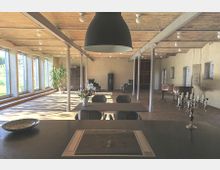 Offener, lichtdurchfluteter Raum mit großen Fensterfronten auf der linken Seite, sichtbaren Ziegelwänden und einer Holzdecke. Der Raum ist mit einem Esstisch samt Stühlen, einem Klavier, einem Kamin sowie Dekorationselementen wie einem Kronleuchter und Pflanzen ausgestattet.