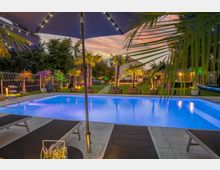 Beleuchteter Garten mit einem rechteckigen Pool, umgeben von Palmen in dekorativen Töpfen und farbigen Lichtern. Im Vordergrund sind Liegestühle und ein Sonnenschirm zu sehen, im Hintergrund eine gepflasterte Wegführung und weitere Sitzbereiche.