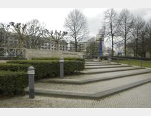 Treppenanlage des Theodor-Heuss-Platzes mit gepflastertem Boden, gesäumt von niedrigen Hecken und Pollerleuchten. Im Hintergrund stehen blattlose Bäume, moderne Gebäude und ein blau-weißer Turm.