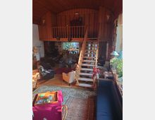 Wohnzimmer mit holzverkleideter Decke und Galerie im oberen Bereich, die über eine hölzerne Treppe mit geschmücktem Geländer erreichbar ist. Das Zimmer ist mit Teppichen, Polstermöbeln, einem Bücherregal und Dekorationen ausgestattet, und große Fenster lassen Tageslicht herein.