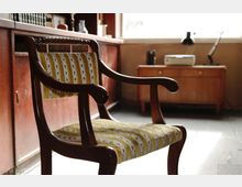 Ein traditioneller Holzstuhl mit gepolsterter Sitz- und Rückenlehne in gelblich-grünem Stoff mit floralen Streifen steht in einem Raum mit Holzmöbeln und einem Sideboard im Hintergrund. Durch ein großes Fenster fällt Tageslicht in die Szene.