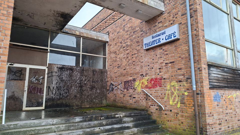 Eingangsbereich eines Backsteingebäudes mit Betonvordach und breiten Stufen, teils mit Graffiti und verwitterten Oberflächen. An der Fassade hängt ein Schild „Restaurant THEATER • CAFÉ“, daneben große Fensterflächen und teils vernagelte Öffnungen.