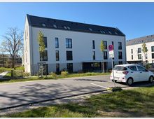 Modernes, dreigeschossiges Wohngebäude mit weißer Fassade, dunklem Satteldach und schmalen Fenstern entlang einer asphaltierten Straße. Im Vordergrund stehen ein Verkehrszeichen, ein geparktes Auto sowie junge Straßenbäume und Grünflächen; daneben sind weitere ähnliche Wohnblöcke zu sehen.