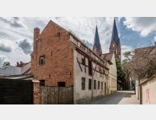 Historische Gasse mit einem Fachwerkhaus und einem angrenzenden Backsteingebäude, im Hintergrund ragen zwei gotische Kirchtürme mit spitzen Dächern in den Himmel. Der Weg ist mit Kopfsteinpflaster ausgelegt, und umliegende Bäume sowie Gebäude säumen die Szene.