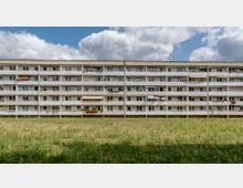 Mehrstöckiges Wohngebäude mit einfachen, identischen Balkons, teilweise dekoriert mit Pflanzen und Markisen. Vor dem Gebäude erstreckt sich eine große, grasbewachsene Fläche, darüber ein Himmel mit weißen Wolken.