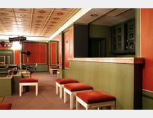 Innenraum eines kleinen Theaters oder Aufführungsraums mit rot-grüner Wand- und Deckenverkleidung, gepolsterten Sitzhockern und Beleuchtungsequipment. Eine Theke aus hellem Material befindet sich rechts im Bild, daneben ist ein Schrank mit Glastüren erkennbar.