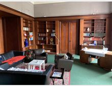 Wohnzimmer oder Bibliothek mit Holzwandregalen, die Bücher und dekorative Objekte enthalten. Der Raum ist mit dunklen Ledersesseln, einem Sofa, einem kleinen weißen Beistelltisch und einem großen Holzschreibtisch auf einem grünen Teppich ausgestattet.