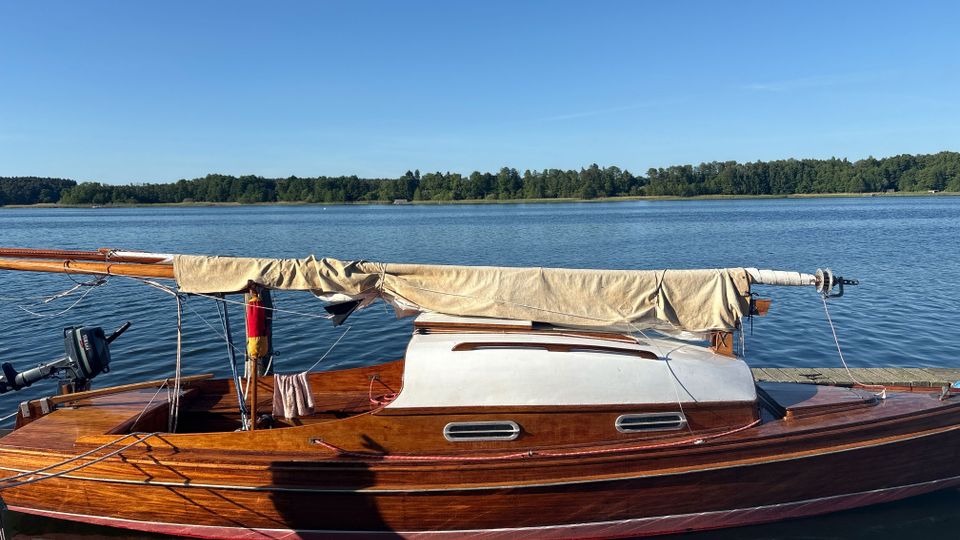 Holzsegelboot mit zusammengebundenem, beige abgedecktem Mast liegt an einem Steg auf einem ruhigen See. Im Hintergrund sind eine bewaldete Uferlinie und ein klarer blauer Himmel zu sehen.