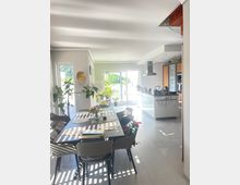 Moderner, heller Ess- und Wohnbereich mit einem großen Esstisch, umgeben von Stühlen, dekoriert mit Platzsets und Blumen. Im Hintergrund eine offene Küche mit weißen Fronten und große Fenster, die zu einer Terrasse mit Pflanzen führen.