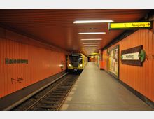 U-Bahn-Station „Halemweg“ mit orangen Wänden, gelben Hinweisschildern und einem gelben Zug, der auf der linken Seite am Gleis hält; rechts sind ein Ausgangsschild und Informationstafeln sichtbar.