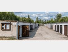 Reihe von flachen Garagen mit Holz- und Metalltoren, die sich entlang eines gepflasterten Weges erstrecken; daneben eine Gebäudevorderseite mit einem Schaukasten und Hinweis. Im Hintergrund sind Bäume und ein wolkiger Himmel zu sehen.