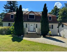 Zweistöckiges Einfamilienhaus mit weißer Fassade und rotem Dach, umgeben von hohen Bäumen und einem gepflegten Garten. Eine halbkreisförmige Treppe mit gepflastertem Weg führt zu einer zentralen weißen Eingangstür, flankiert von zwei hohen Zypressen.