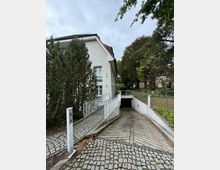 Wohngebäude mit grauer Fassade und rotem Satteldach, daneben eine schräge Rampe mit Geländer, die zum Gebäude führt. Im Vordergrund befindet sich eine gepflasterte Einfahrt, die zu einer Tiefgarage hinunterführt, umgeben von Bäumen und einem Zaun.