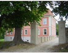Ansicht eines historischen Gebäudes mit rosa Fassade und weißen Fensterrahmen, umgeben von Bäumen. Im Vordergrund befinden sich ein großer Baum und zwei steinerne Eingangspfeiler, die einen gepflasterten Weg zum Gebäude führen.