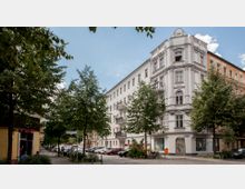 Ecke einer städtischen Wohnstraße mit einem mehrstöckigen Altbau mit verzierten Fassadenelementen. Vor dem Gebäude befinden sich Bäume, parkende Autos und ein Gehweg mit niedrigen Pollern.