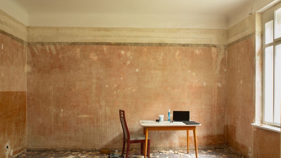 Ein leerer, renovierungsbedürftiger Innenraum mit abblätternden Wänden und bröckeligem Boden, in dessen Mitte ein kleiner Tisch mit Stuhl und Laptop steht. Rechts lässt ein großes Fenster Tageslicht in den Raum.