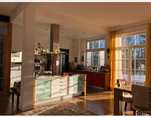 Helles Wohnküchen-Interieur mit großen Fenstern und gelben Vorhängen, durch die Sonnenlicht einfällt. Die Küche verfügt über eine Kochinsel mit hellen Schubladen, roten Hängeschränken und einem Edelstahl-Dunstabzug; im Vordergrund ein Holztisch und ein Teppich auf Parkettboden.