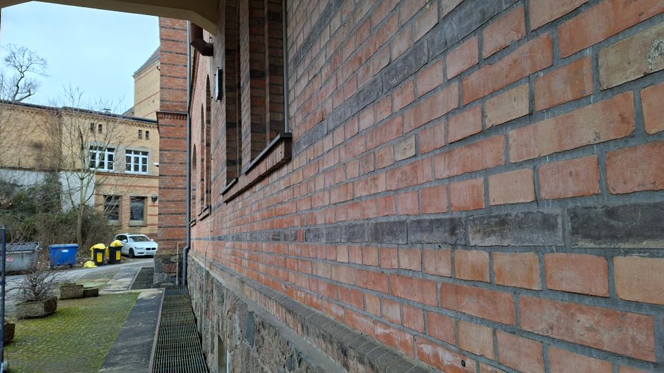 Schmaler Außenweg entlang einer langen Ziegelsteinfassade mit Rundbögen und Sockel aus Naturstein, teilweise mit Metallgitter-Abflussrinne am Boden. Im Hintergrund stehen weitere Backsteingebäude sowie Mülltonnen und ein geparktes Auto in einem Hofbereich.