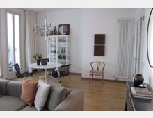 Modernes Wohnzimmer mit Holzboden, weiß gestrichenen Wänden und großen Fenstertüren mit Vorhängen. Im Raum befinden sich ein graues Sofa mit Kissen, ein runder weißer Esstisch mit schwarzen Stühlen, eine Glasvitrine mit Dekorationsgegenständen sowie ein minimalistischer Kronleuchter.