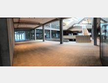 Offener Innenraum eines modernen Bürogebäudes mit einem großen Holzfußboden, sichtbaren Betonelementen und Glaswänden. Im Hintergrund führt eine breite Treppe zu einer Empore, während die Decke große runde Lichtquellen aufweist.