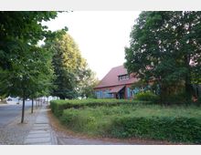 Straßenszene mit einem gepflasterten Gehweg und Bäumen entlang der Straße. Rechts steht ein Backsteinhaus mit rotem Satteldach, blauen Fensterläden und einem Vorgarten, umgeben von einer Hecke und Bäumen.