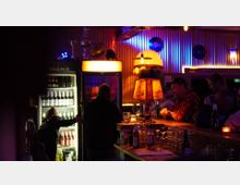 Ein Innenraum einer Bar mit dunkler, stimmungsvoller Beleuchtung. Im Vordergrund steht eine Theke mit Gläsern, Flaschen und einer warm beleuchteten Tischlampe; im Hintergrund sind ein offener Kühlschrank mit Getränken und Menschen erkennbar.